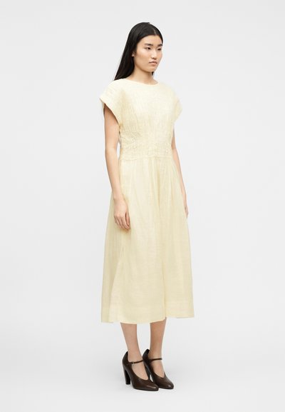 Γυναίκα που στέκεται πλάγια φορώντας ένα μίντι φόρεμα με υφή σε ανοιχτό κίτρινο χρώμα με κοντά μανίκια και σκούρα καφέ γόβες Mary Jane με ψηλό τακούνι.