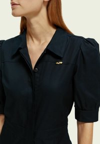 Vrouw in een zwarte gekraagde jurk met pofmouwen en een kleine gouden broche op de linkerborst.