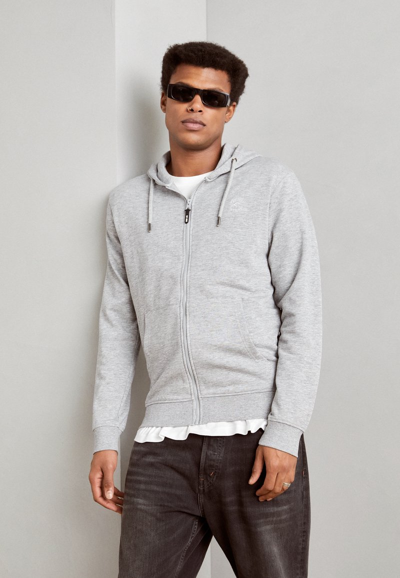 Gray zip-up hoodie with a drawstring hood, pockets, and ribbed cuffs, layered over a white shirt. Model wearing dark jeans and sunglasses.