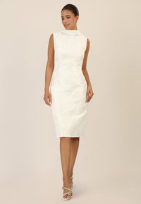 White sleeveless dress with floral textured pattern, fitted shape, and draped neckline. Paired with white strappy sandals.