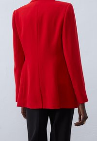 Veste rouge ajustée, avec manches longues, une fente unique au dos, et un tissu lisse. Portée sur un pantalon noir.