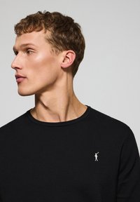 Suéter negro de punto con diseño ajustado. Presenta un pequeño logo blanco en el lado izquierdo del pecho. Textura suave y silueta clásica.