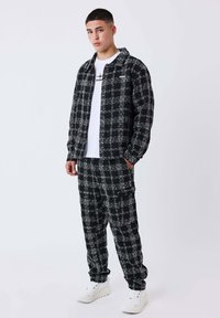 Giovane uomo che indossa una giacca e pantaloni a scacchi bianco e nero, una maglietta grafica bianca e sneakers bianche, in piedi di fronte a uno sfondo neutro.