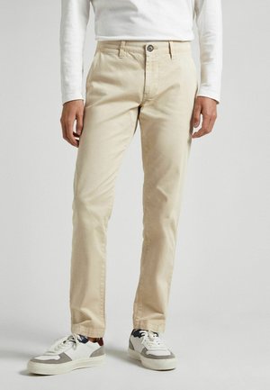 Man wearing beige pants, white long-sleeve shirt, and white sneakers with gray and navy blue details, standing against a plain background.