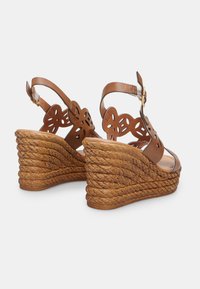 Sandales compensées marron avec un design floral découpé, une lanière de cheville ajustable et une plateforme tissée texturée. Le talon mesure environ 8 cm de hauteur.