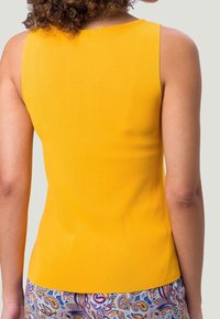 Person, der bærer en ærmeløs gul top og bukser med paisley-mønster, vist fra ryggen mod en ensfarvet baggrund.