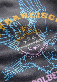 Šedá tkanina se grafickým designem orla s modrými křídly, zlatým a růžovým textem a štítem se hvězdami, na kterém je vytištěno "California".