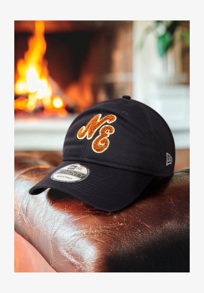 Mustaline must reguleeritav müts, millel on tekstuuriga oranž ja kreemikas lõngaga bordüüritud "NE" logo, asetatud pruuni nahapinna peale.