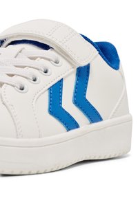 Baskets blanches avec des accents bleus, dotées d'une sangle en velcro, d'un dessus texturé et de deux bandes zigzag bleues sur les côtés. Semelle en caoutchouc.