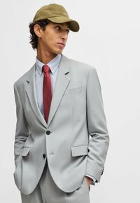 Light gray suit jacket with two front pockets, black buttons, paired with a light blue shirt and a red dotted tie. Olive green cap worn casually.