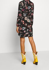 Frau in einem schwarzen Blumenkleid und gelben Cowboy-Stiefeln, die vor einem schlichten hellen Hintergrund steht und abgewandt ist.