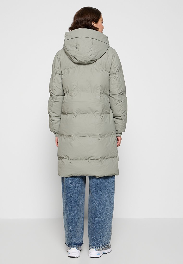 Grau gepolsterter Mantel mit Kapuze, ausgestattet mit einem gesteppten Design und einer langen Silhouette, kombiniert mit blauen weiten Jeans und weißen Sneakers.