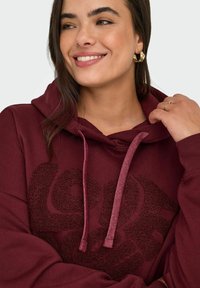 Burgundy huppari, jossa on teksturoitu "Rakkaus"-design, varustettu nyörillä ja ribbausmanjeteilla. Tasainen kangas ja rento malli.