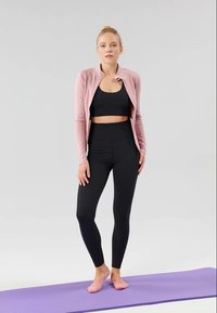 Crne sportske legginsice i crni crop top nosi se ispod svijetlo ružičaste jakne na patent zatvarač. Osoba stoji na ljubičastom jogi prostirku.