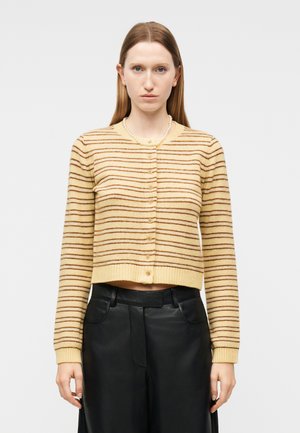 Γυναίκα που φοράει μπεζ και καφέ ριγέ πλεκτή ζακέτα με κουμπιά και μακριά μανίκια, σε συνδυασμό με μαύρο ψηλόμεσο παντελόνι, μπροστά σε απλό φόντο.