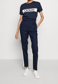 Marineblå t-shirt med hvid "HUGO" logo, parret med marineblå bukser med hvide sidelister. Modellen har hvide sneakers på. Glat stof, afslappet pasform.