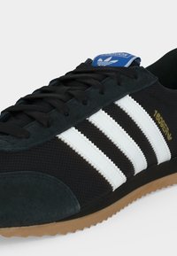 Μαύρο αθλητικό παπούτσι Adidas με λευκές ρίγες, καουτσούκ σόλα, μαύρα κορδόνια και μπλε λογότυπο Adidas στη γλώσσα, αριθμός μοντέλου 16099ER σε χρυσό στο πλάι.