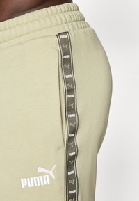 Un pantalon de survêtement vert olive avec une taille élastique, présentant une bande latérale avec des logos Puma blancs répétés et un logo brodé sur le devant.