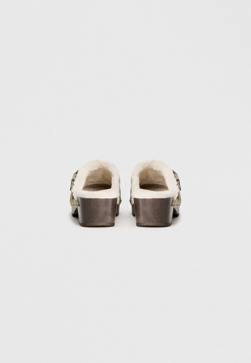 Clogs mit einem Holzsockel, weißem Fellfutter und einem verstellbaren Riemen mit metallischen Akzenten; Rückansicht mit einem abgerundeten, offenen Design.