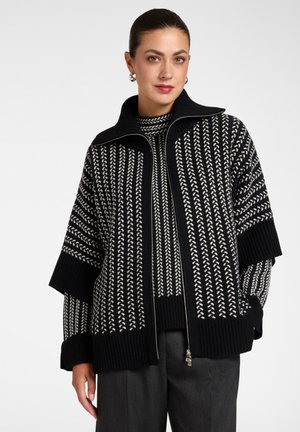 Woman with dark hair in a bun wearing black and white patterned knit sweater with a high collar and zip, paired with dark pants.