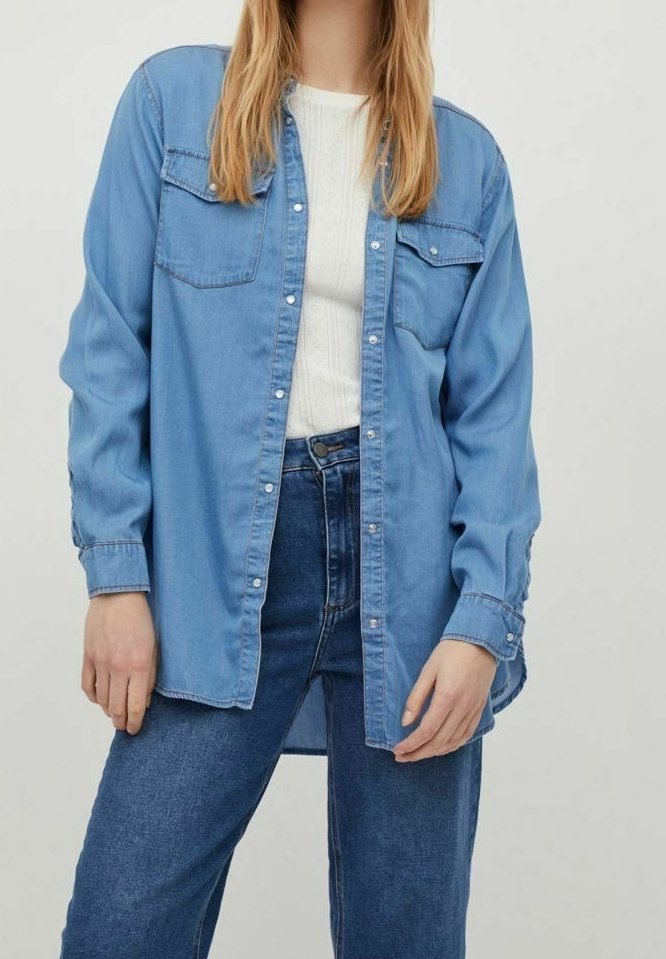 Chemise en denim bleu clair avec deux poches avant, fermeture par boutons, manches longues retroussées aux poignets, et coupe décontractée. Associée à un jean bleu.