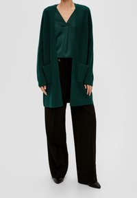 Cardigan verde scuro realizzato in maglia testurizzata, con maniche lunghe, apertura frontale e due grandi tasche anteriori. Abbinato a una blusa teal e a pantaloni neri.