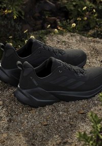Zwarte Adidas Terrex trail hardloopschoenen geplaatst op grind met groene planten en kleine gele bloemen op de achtergrond.