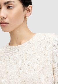 Elegant cremetop med indviklet perle- og palietbroderi, der har en rund halsudskæring og strukturerede mønstre. Diskrete guldringe som øreringe.