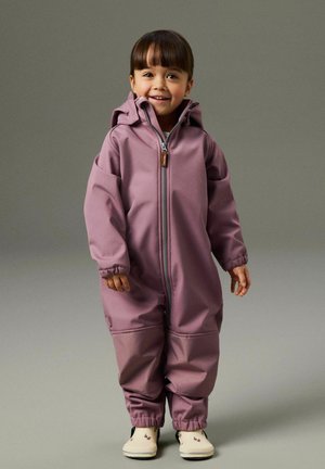 Uśmiechnięte dziecko w różowofioletowym kombinezonie z kapturem i zamkiem z przodu, noszące beżowe buty z wzorem wiśni, stojące na szarym tle.