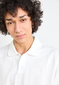 Polo shirt blanc en tissu texturé, arborant un logo brodé subtil sur la poitrine gauche, et un col classique avec une ouverture à bouton.