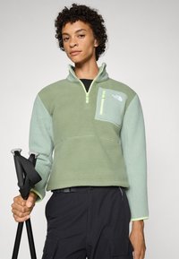 Fleece pullover in groentinten met een halve rits, een ritszak op de borst en contrasterende mouwen. Bevat trekkingstokken in de hand.