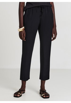 Femme portant un pantalon noir court avec une taille à cordon, des sandales noires avec des accents dorés et des bracelets blancs et dorés au poignet gauche.