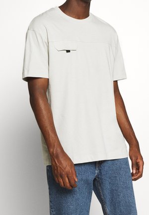 Mann trägt ein hellgraues T-Shirt mit kurzen Ärmeln und Brusttasche sowie blaue Jeans, steht vor einfachem weißem Hintergrund.