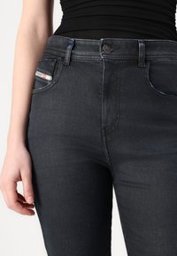 Jeans en denim noir avec un design ajusté, légèrement délavés, et une petite étiquette logo sur la poche avant. Fermeture à bouton à la taille.