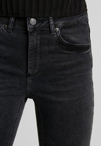 Jeans de denim negro con una forma ajustada, que cuentan con un bolsillo frontal y un cierre de botón metálico. La tela presenta una textura suave con un ligero desvanecimiento.