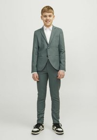 Un costume vert ardoise avec une veste à un seul bouton, un pantalon ajusté, et une chemise blanche à col, associé à des baskets noires et blanches.