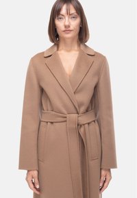 Cappotto lungo in lana marrone con colletto a scialle, vita con cintura e tasche laterali; presenta una texture morbida e una vestibilità sartoriale.