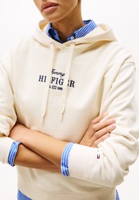 Krémová mikina s kapucňou a vyšívaným logom "Tommy Hilfiger". Preložená s modrou pruhovanou košeľou, s kontrastnými manžetami.