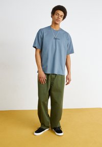Modrá bavlněná tričko s krátkými rukávy a logem "Dickies". Olivově zelené kalhoty s širokými nohavicemi, které jsou kombinovány s černými teniskami s bílou podrážkou.