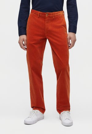 Homme portant un pantalon en velours côtelé orange vif, une chemise à manches longues bleu marine et des baskets blanches, debout les mains le long du corps.