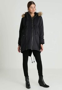 Woman wearing a black winter parka with fur-trimmed hood, black leggings, and black ankle boots, standing against a plain light background.