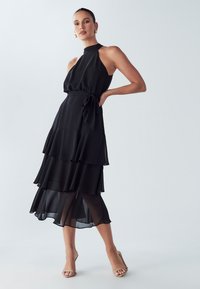 Musta hihaton mekko, jossa on korkea kaulus ja kerroksittainen röyhelösomistus. Valmistettu kevyestä kankaasta, siinä on vyötärösidos ja läpikuultavat kerrokset.