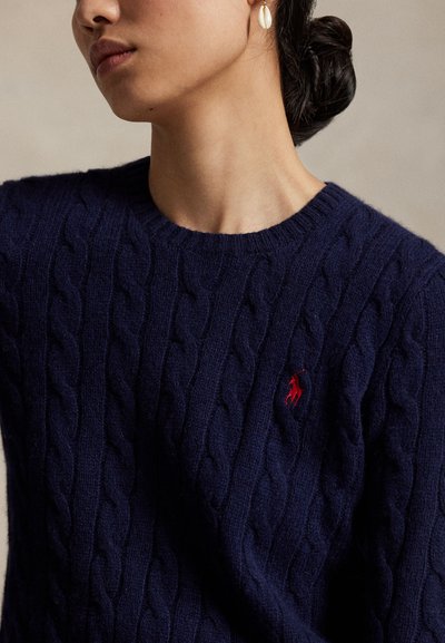 Femeie cu un pulover bleumarin tricotat cu model de cablu, cu logo roșu brodat și cu o singură cerceață în formă de scoică, părul prins într-un coc jos.