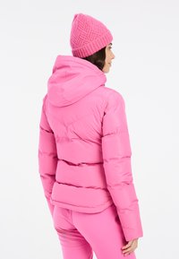Giacca a vento rosa con cappuccio, caratterizzata da cuciture orizzontali e una silhouette aderente. Indossata con un berretto rosa a trama coordinato.