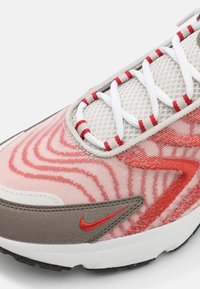 Gros plan d'une basket Nike beige et rouge avec des lacets blancs et une tige en mesh à motifs sur fond blanc.