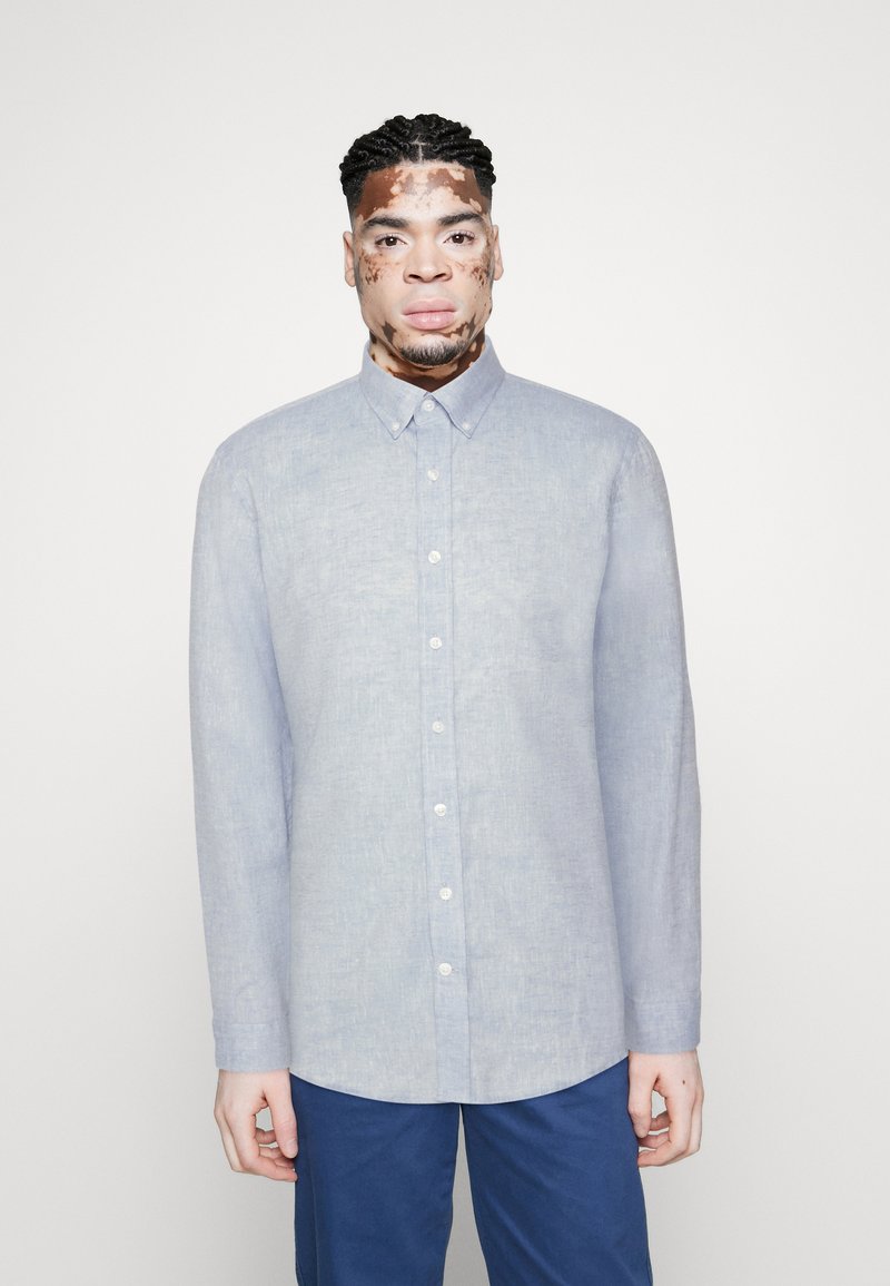 Cămașă button-up de culoare bleu deschis, realizată din in, cu guler clasic, mâneci lungi și un tiv ușor curbat.