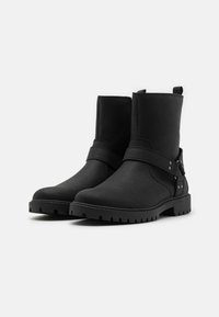 Botas de tobillo negras de cuero con superficie texturizada, detalle de correa, acentos metálicos y suelas de goma resistentes con un patrón de tacos.