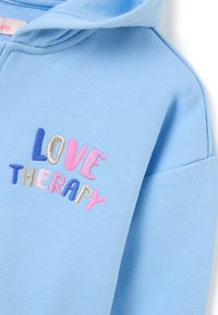 Hellblaues Hoodie mit weicher Textur und großer "LOVE THERAPY" Stickerei in rosa, blau und gold. Einfaches Design und lässige Passform.