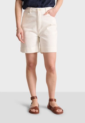 Shorts en coton couleur crème avec une taille haute, un ourlet roulé et des poches latérales. Associés à des sandales à lanières marron.
