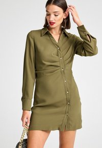 Olivgrünes Hemdkleid aus weichem Material mit Kragen und durchgehender Knopfleiste. Verfügt über Faltendetails und Schlitze an den Ärmeln. Knöpfe in Goldoptik.
